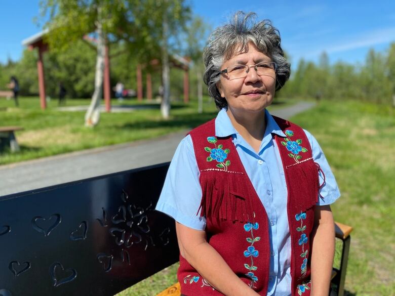 A woman in glasses and a beaded vest sits outside in the summer.