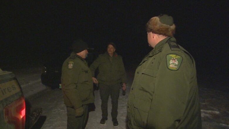 Three men in green uniforms with crests on the shoulder stand near vehicles on a dark road.