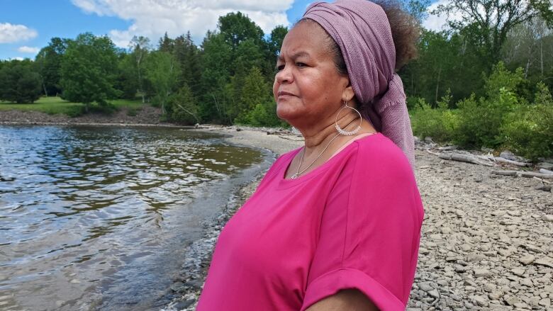 A Black woman stands on the shore of a river, looking out at the water, with her curly dark hair pulled back in a mauve-coloured scarf and she wears a pink t-shirt. 