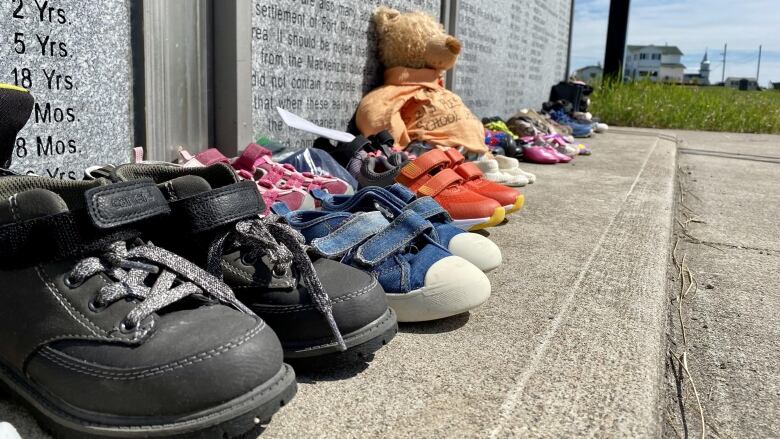 Shoes and a teddy bear in front of a large plaque bearing many names.