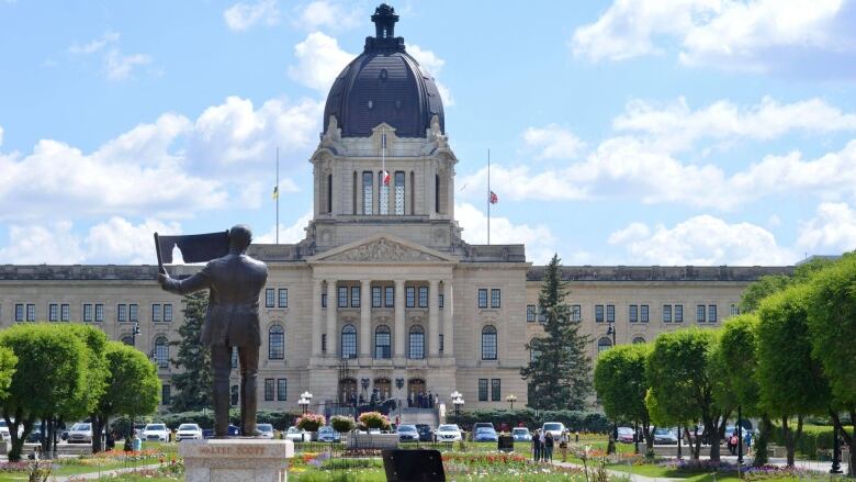 A blue sky with white puffy clouds hang above a government building with a tower. The building stands behind a parking lot filled with vehicles and a garden of flowers of red, purple and yellow. There's a statue erected in the garden. Trees line the edge of the green space.