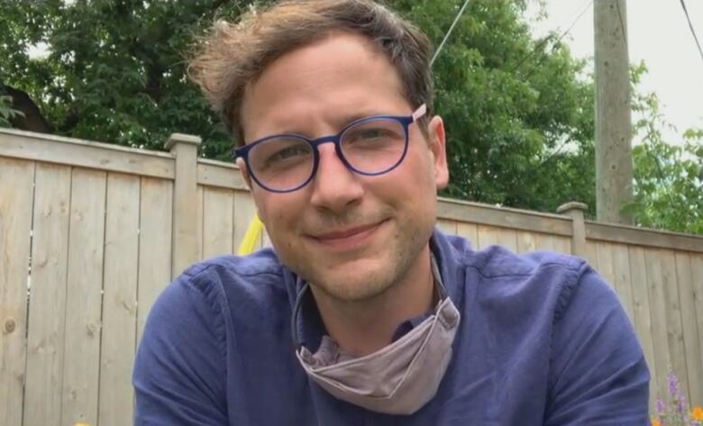 Man with short brown hair, thin blue glasses and a mask wearing a blue shirt.