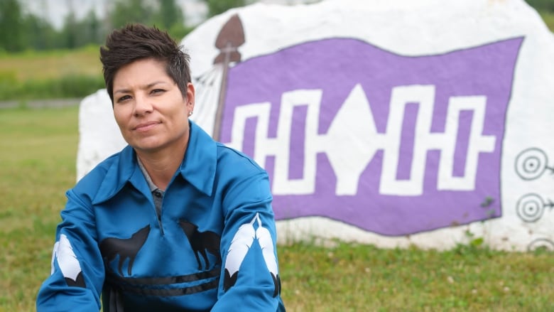 A woman wearing a ribbon shirt sits on the grass in front of a large rock painted with the Haudenosaunee Confederacy flag.