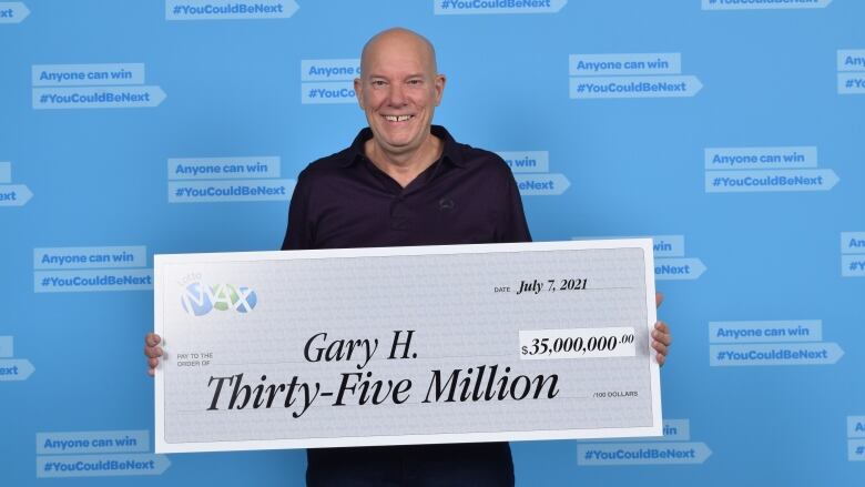 A middle-aged white man stands holding a novelty lottery cheque for thirty-five million that says Gary H. on it. He wears a black shirt and a huge smile.