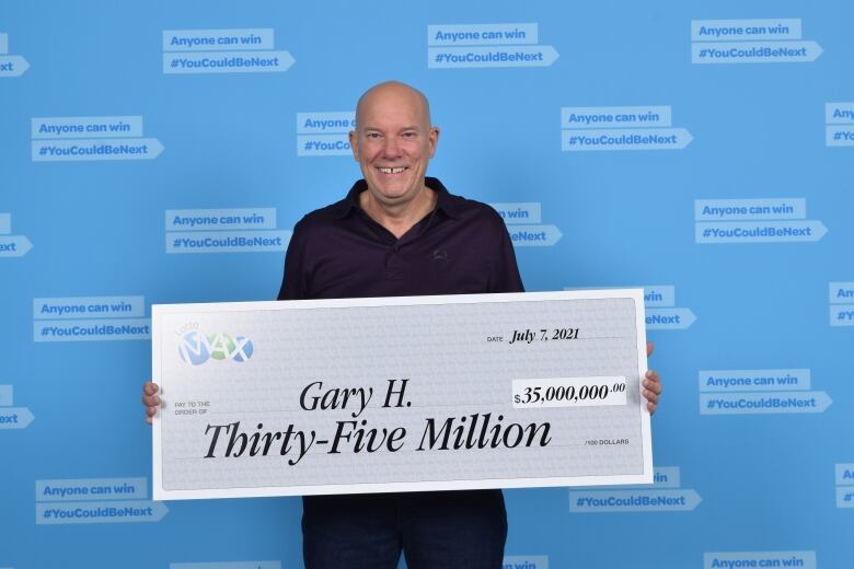 A middle-aged white man stands holding a novelty lottery cheque for thirty-five million that says Gary H. on it. He wears a black shirt and a huge smile.