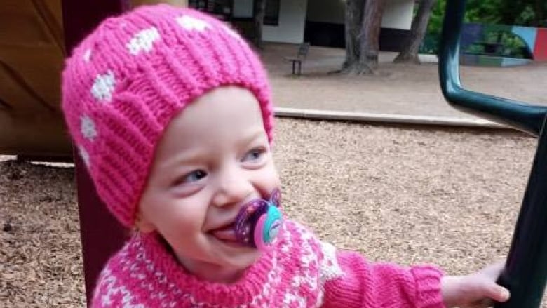 A toddler sucking a soother plays in a playground with wood chips on the ground. She is wearing a pink toque and pink sweater and is smiling.