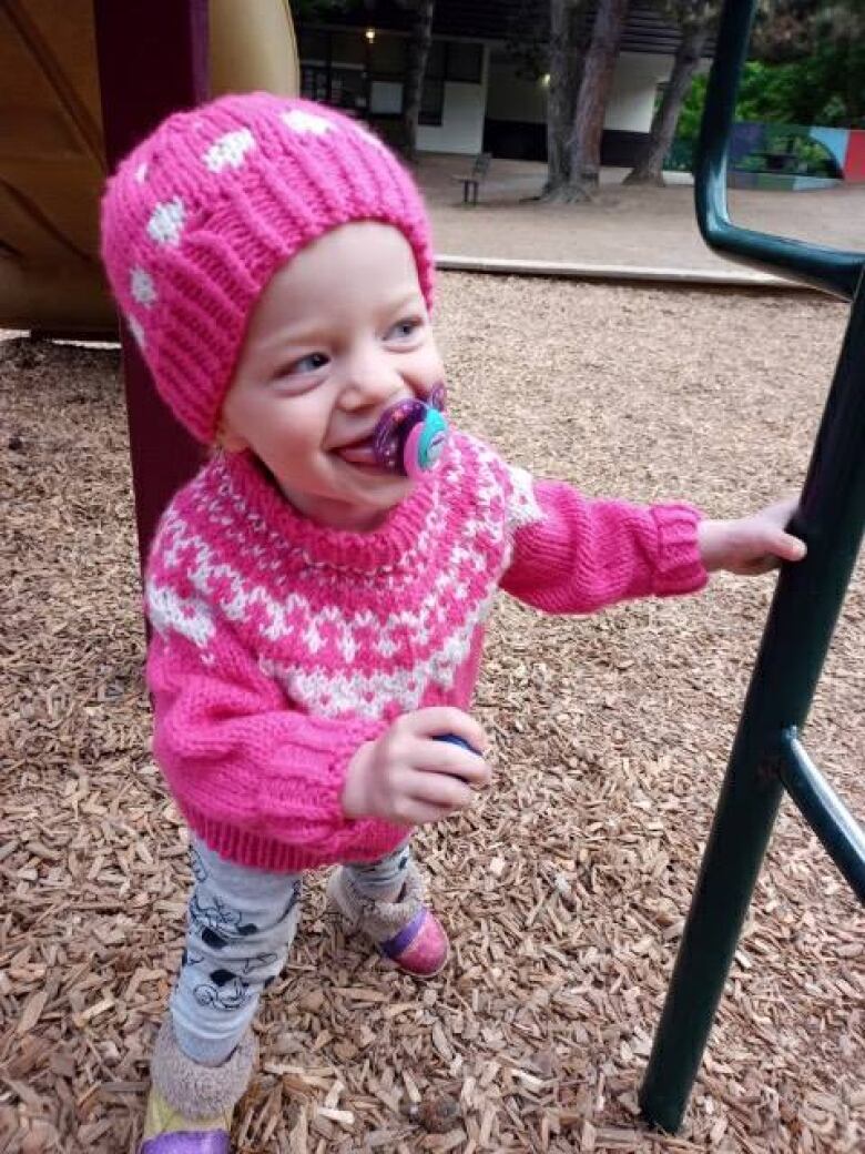 A toddler sucking a soother plays in a playground with wood chips on the ground. She is wearing a pink toque and pink sweater and is smiling.