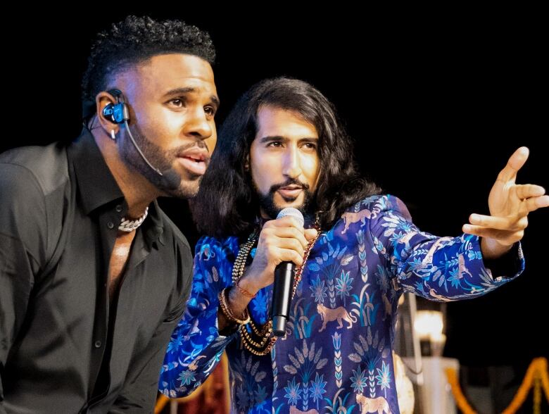 Two black-haired men sing into microphones, with the one on the far left wearing a dark shirt and jacket, and the one at right dressed in a bright blue Indian suit and wearing necklaces.