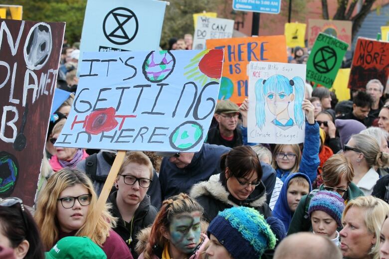 A large crowd stands with signs with climate-change related slogans. One says It's getting hot in here, another says there is no planet B. 