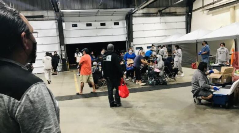 A number of people gathered in an airport hanger, with some emergency supplies in the background.