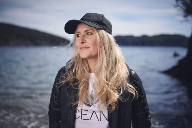 A woman wearing a black jacket and black baseball hat standing in front of a lake. 