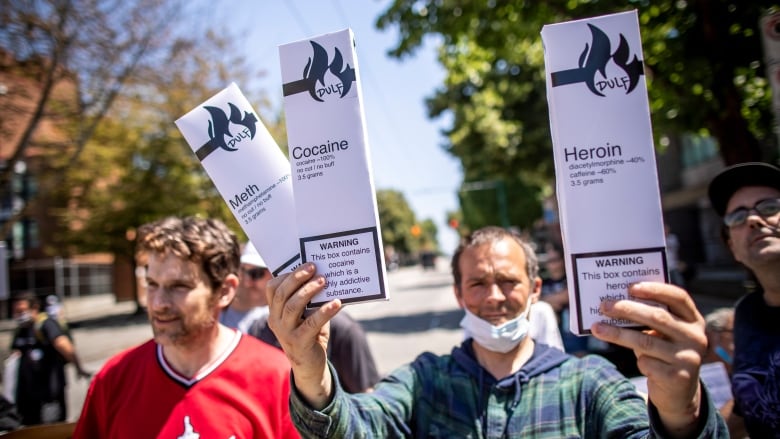 A man wearing a mask around his chin holds up three boxes, styled like cigarette cartons. They are marked 'Meth', 'Cocaine' and 'Heroin'. The man is surrounded by other people.