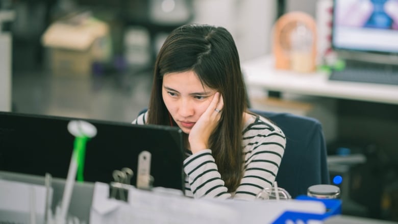 Young woman sitting at a computer in an open-concept office, with a hand on her face, looking tired. 