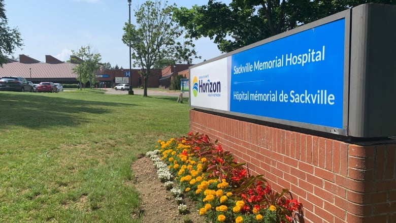 A blue and white sign sits on a brick base on the lawn in front of a hospital building, visible in the distance. There are flowers planted in front of the sign and the sign says 