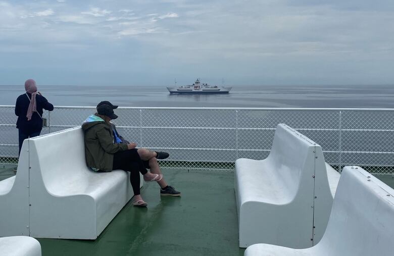 Three passengers stand on the green-painted deck of a passenger ferry in the middle of a stretch of blue ocean.