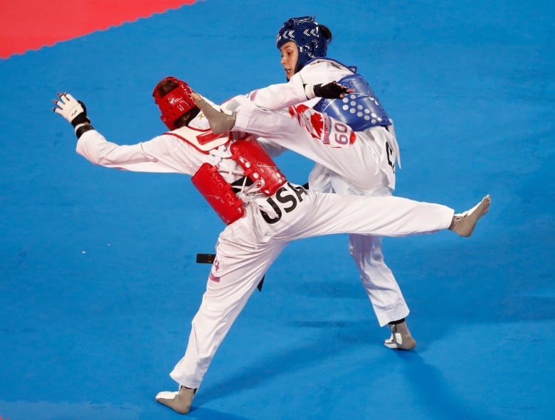 Two people in taekowndo gear kick as they spar on a blue mat.