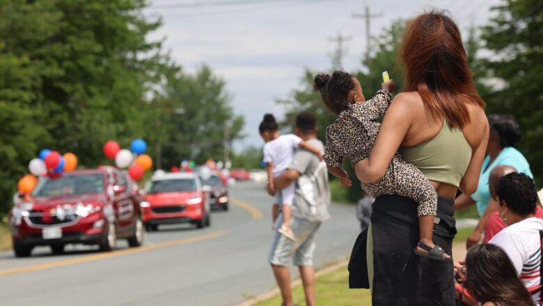 A person holding a baby looking on at a parade procession