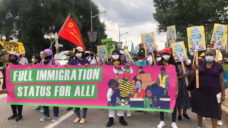 A group of people standing in a street hold placards and march behind a sign reading 