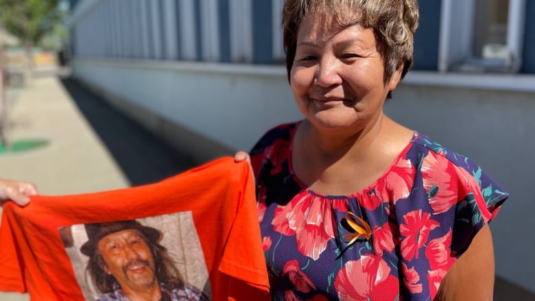 A woman holds a T-shirt emblazoned with the picture of a smiling man with long hair and a mustache wearing a hat.