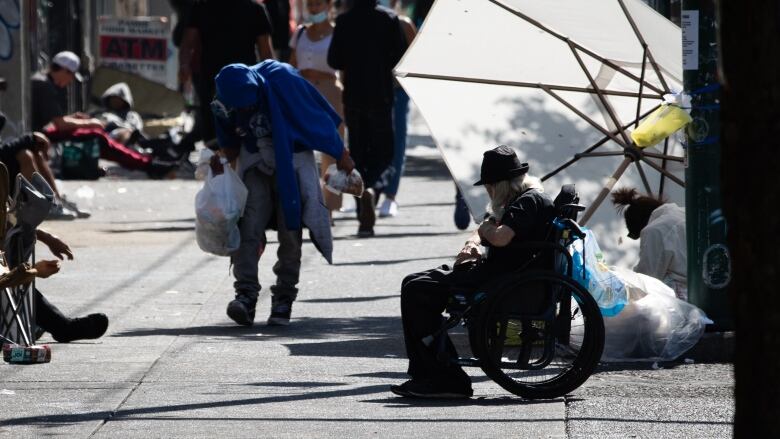 A large umbrella blocks the sun as people sit on the sidewalk.