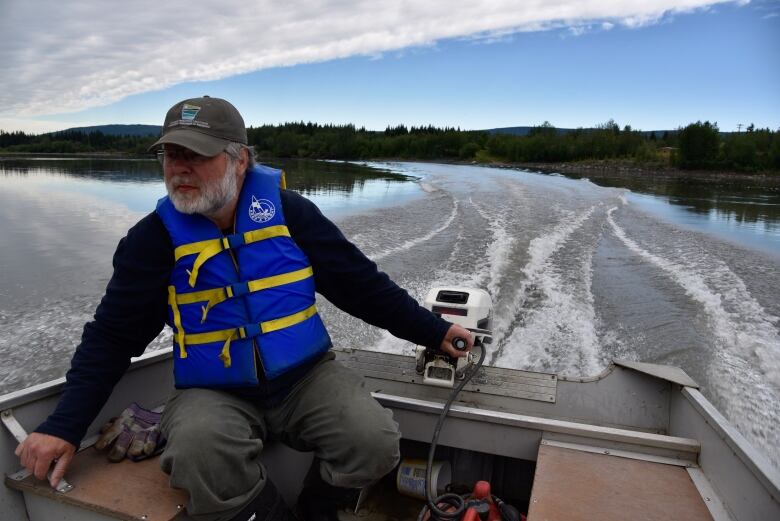 A man with a white beard and wearing a safety vest is in a small motorboat crossing a river.