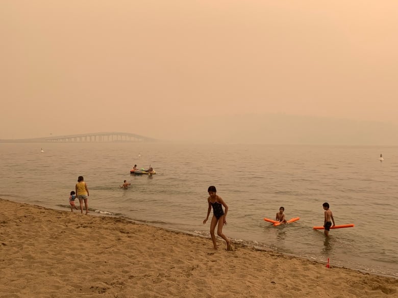 Children play on the Hot Sands Beach in downtown Kelowna, B.C., near the Okanagan Lake with low visibility of William R. Bennett Bridge amid heavy wildfire smoke on July 30, 2021.