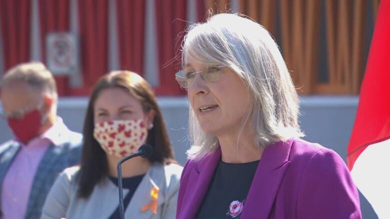 A woman with shoulder-length grey hair and purple blazer speaks outside at a microphone with people standing beside her.