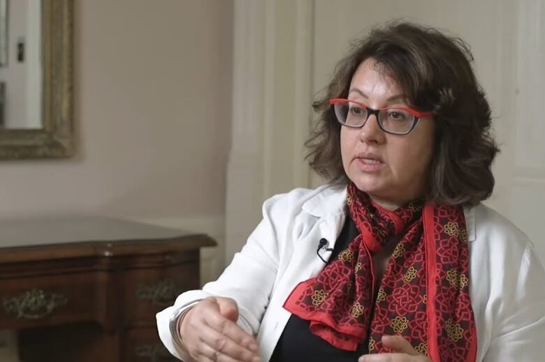 A woman wearing glasses, a white blazer and a red scarf speaks.