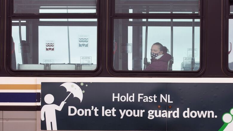 A lone woman sits in a bus, which has an ad on the side reading Hold Fast N.L., Don't let your guard down.