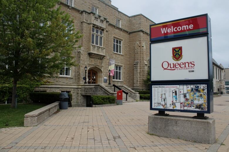 A sign says 'Welcome - Queen's University' in front of a stone building.