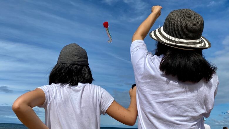 Two people wearing hats and flying a kite are seen from behind on a P.E.I. beach. 