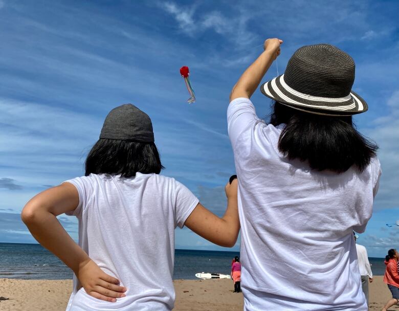 Two people wearing hats and flying a kite are seen from behind on a P.E.I. beach. 