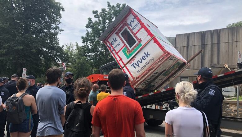 People watch as a white wooden shelter is loaded onto a tow truck