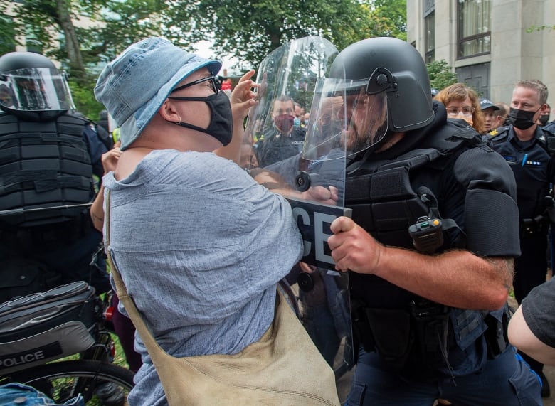 A person wearing a face mask faces off against a police officer in riot gear.