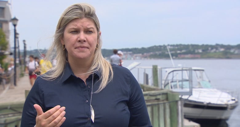 A woman in a blue jacket speaks in front of boats and water.