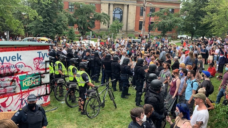 Halifax police in riot gear and with bicycles form a wall between a shelter and protestors outside the old library on Spring Garden Road in August 2021. 