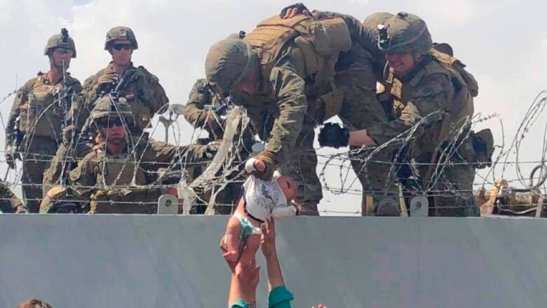 Troops in camoflage reach down over barbed wire to grab a baby being passed up to them