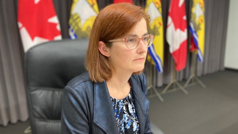 The profile of a woman with red hair and glasses, wearing a black leather jacket, seated in a black leather chair, with Canada and New Brunswick flags in the background.