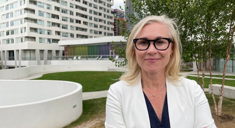 A woman with blond hair, wearing black rimmed glasses and a white blazer, poses in a park near a condo tower.