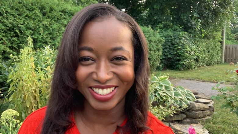 A smiling Black woman with long dark hair in a garden wearing a red blouse