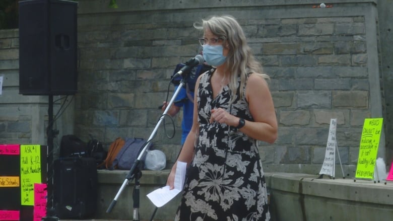 A woman with long grey hair and glasses wears a blue medical mask as she speaks into a microphone at a protest.