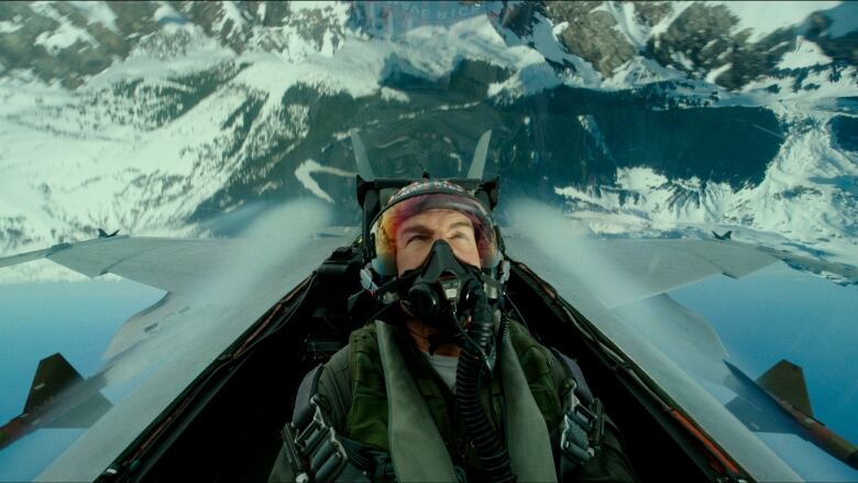 A man in pilot's gear sits in a cockpit surrounded by snowy mountains.