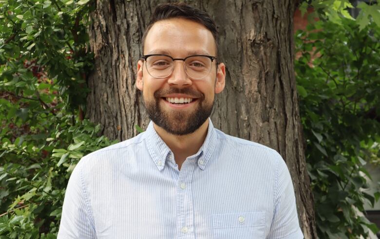 A man with brown hair and a brown beard wearing glasses and a light blue shirt standing in front of a tree