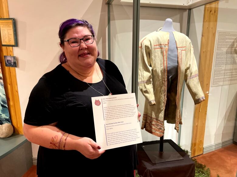 A woman holds a plaque for an Innu coat. A painted caribou hide coat is behind her.