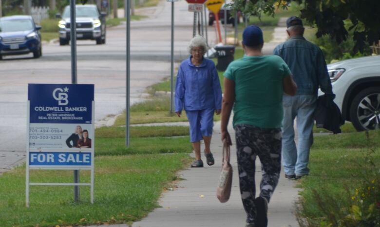 Several people walk on a sidewalk in a residential neighbourhood of North Bay, past a for sale sign. 