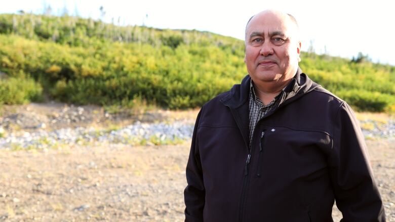 A man in a black jacket stands in front of a hill. 