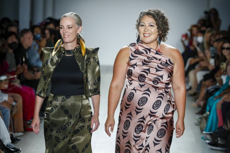 Two woman wearing high fashion clothing with indigenous design walk on a runway. 