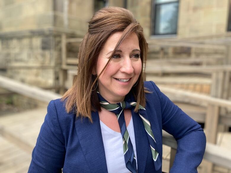 A woman with brown hair in a blue jacket is smiling in front of a stone building.