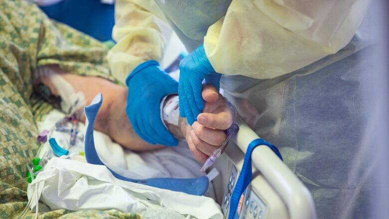 A closeup of a health-care professional wearing a yellow gown and blue medical gloves tending to the IV in the arm of a patient lying in a hospital bed.