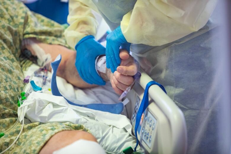 A closeup of a health-care professional wearing a yellow gown and blue medical gloves tending to the IV in the arm of a patient lying in a hospital bed.